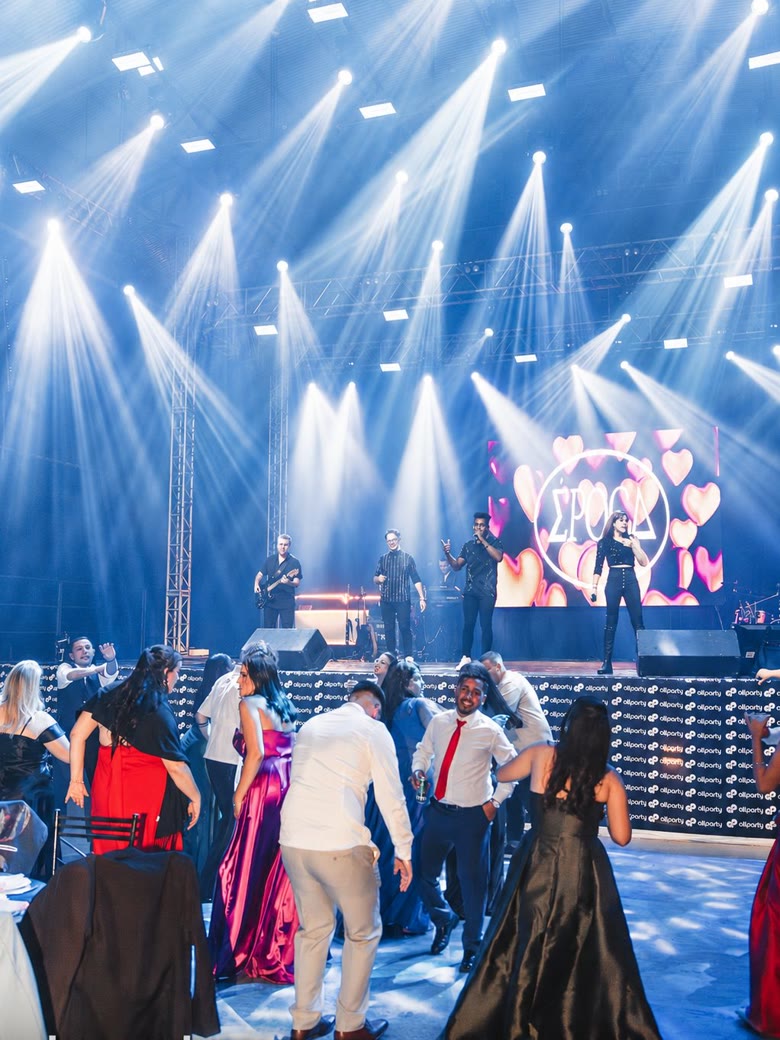 Convidados em trajes de gala dançando em frente à banda, com telão de LED animado ao fundo