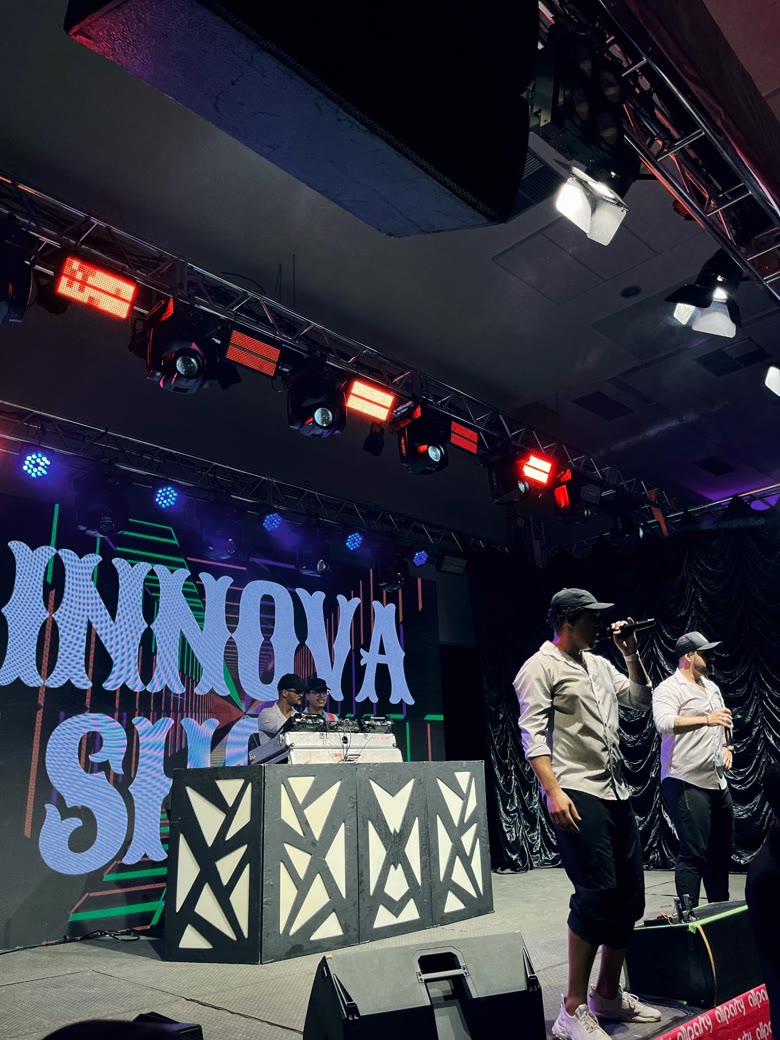 Apresentadores no palco do Innova Show, com telão de LED e DJ ao fundo
